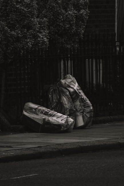 Several large black rubbish bags, made of plastic and slightly crumpled, are piled on the edge of a paved sidewalk near the curb in an outdoor urban setting. The bags appear filled with discarded items and are leaning against a black metal fence with vertical bars, which extends across the background. Behind the fence, dense leafy greenery is visible, suggesting a garden or hedgerow area, with some branches and leaves partially illuminated by natural light. The scene is captured during daytime with diffuse lighting, and the bags contrast against the dark color of the fence and the pavement. The overall environment indicates a private space or the boundary of a residential property, possibly prepared for collection or disposal by private waste services such as Waste Collection Watford, which specializes in rubbish removal. The image portrays a typical scenario of domestic waste awaiting clearance in an indoor-outdoor transitional space relevant to private rubbish collection services involved in on-site clearance or alternative waste handling methods.