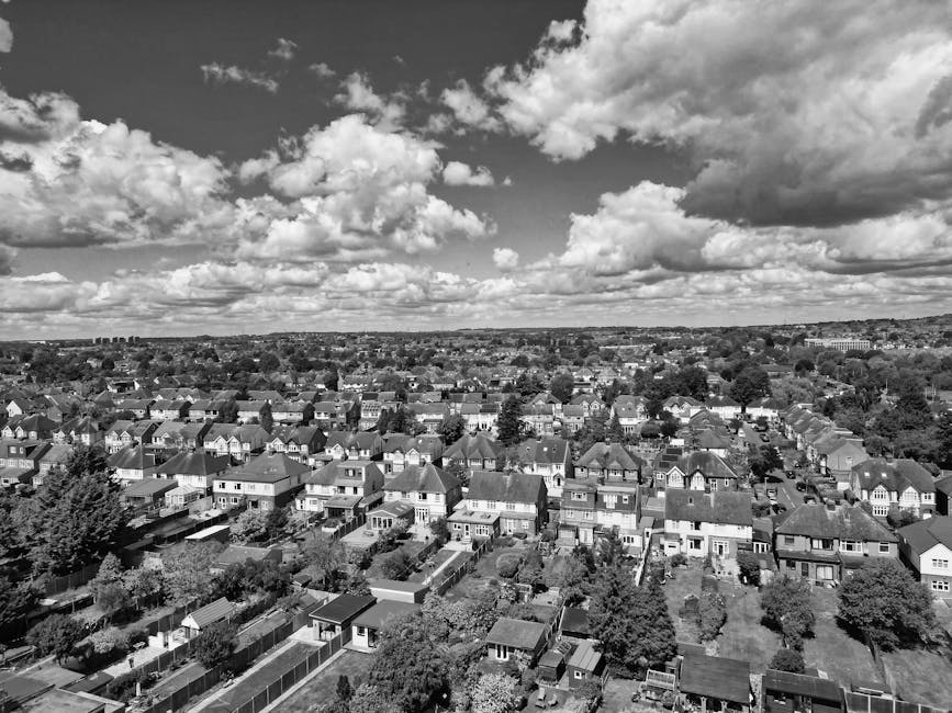 A black and white aerial photograph showing a dense residential neighborhood with rows of closely packed houses and detached properties, each featuring varied pitched roofs, some with chimneys, and small front and back gardens. The houses are arranged in a grid pattern with narrow streets and alleyways separating them. In the foreground, there are multiple small garden sheds and neatly maintained yards, indicating typical domestic waste management areas. The image captures a broad view of the suburban landscape under a sky filled with large, cumulus clouds, creating a textured contrast against the orderly housing layout. The scene reflects typical urban sprawl, with an emphasis on private property and on-site waste disposal methods, consistent with independent rubbish clearance services. This urban environment underscores the importance of professional waste collection companies such as Waste Collection Watford, which offer alternative waste handling options for residential areas to support efficient rubbish removal and sanitation.