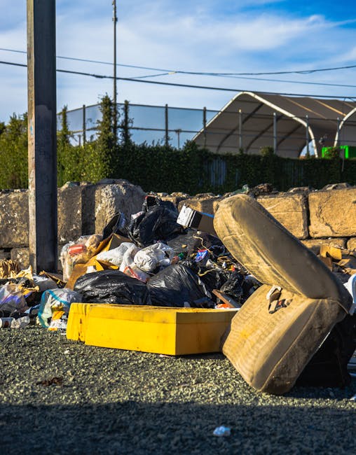 Several large black rubbish bags, made of plastic and slightly crumpled, are piled on the edge of a paved sidewalk near the curb in an outdoor urban setting. The bags appear filled with discarded items and are leaning against a black metal fence with vertical bars, which extends across the background. Behind the fence, dense leafy greenery is visible, suggesting a garden or hedgerow area, with some branches and leaves partially illuminated by natural light. The scene is captured during daytime with diffuse lighting, and the bags contrast against the dark color of the fence and the pavement. The overall environment indicates a private space or the boundary of a residential property, possibly prepared for collection or disposal by private waste services such as Waste Collection Watford, which specializes in rubbish removal. The image portrays a typical scenario of domestic waste awaiting clearance in an indoor-outdoor transitional space relevant to private rubbish collection services involved in on-site clearance or alternative waste handling methods.