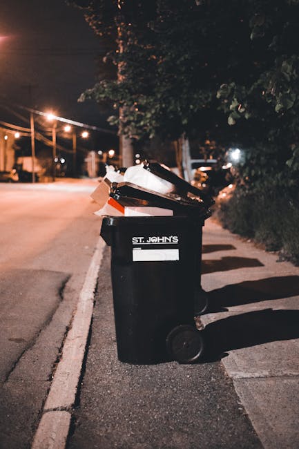 A black wheeled rubbish bin positioned on a paved sidewalk next to a street at night. The bin has a white label with the text 'ST. JOHN'S' on the front and appears to be filled with mixed waste, including cardboard and plastic items, some of which are slightly protruding from the open lid. The surrounding environment includes illuminated streetlights, trees, and residential buildings in the background, with shadows cast on the pavement. The scene reflects an urban setting suitable for private or independent waste collection services, such as those offered by Waste Collection Watford, and illustrates typical refuse disposal activity outside a residence during evening hours.