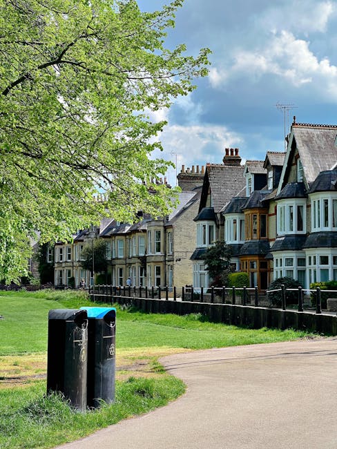 A pair of black recycling bins positioned on a grassy verge beside a residential pavement during daylight hours. The bins are made of plastic with a slightly curved design and are equipped with locked lids, one with a blue lid and the other with a black lid, illustrating standard waste collection containers used for general or recyclable rubbish. Behind the bins, there is a row of traditional terraced houses with bay windows, slate roofs, and brick chimney stacks, typical of UK suburban architecture, set against a backdrop of a partly cloudy sky. A wooden fence runs along the front of the houses, separating the properties from the green area, which features a large deciduous tree with fresh green leaves indicating a spring or early summer scene. The environment appears clean and well-maintained, with no visible litter, and the scene suggests an area serviced by local waste collection providers such as Waste Collection Watford, involved in routine rubbish and recycling management, potentially including private or independent collection services tailored to residential communities.